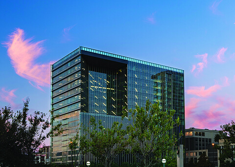 Glass office building with a large square cutout in the facade, set against a pink sunset and trees.