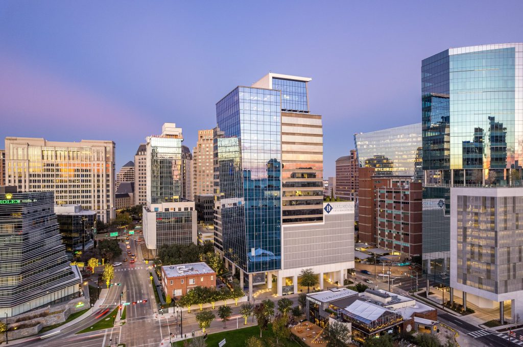 Aerial view of office towers in Dallas’ Harwood District at dusk, with glass high-rises lit against a pink and purple sky above tree-lined streets.